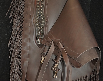 May include: Brown leather chaps with fringe and decorative stitching. The chaps have a belt loop and a buckle closure.