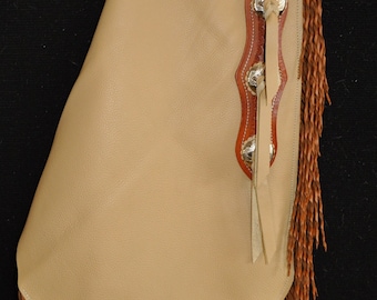 May include: Tan leather chaps with brown fringe and silver conchos. The chaps have a belt loop and a decorative top flap.
