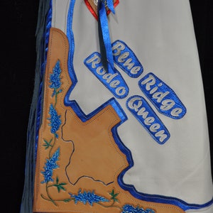 May include: White leather chaps with blue fringe and blue stitching. The chaps feature a brown leather Texas outline with bluebonnets and the text "Blue Ridge Rodeo Queen".