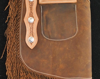 May include: Brown leather chaps with decorative floral tooling and silver-tone conchos. The chaps feature a pocket, fringe along the side, and white stitching. A tag with an American flag is visible.