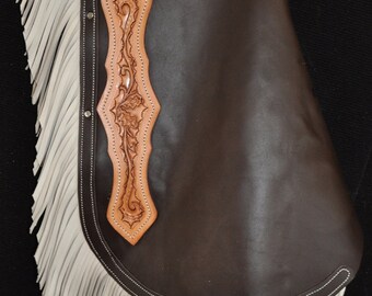 May include: Brown leather chaps with white fringe and intricate tooled designs. The chaps have a belt loop and a decorative strap with a buckle.