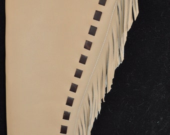 Op de afbeelding: Tan lederen chaps met bruine accenten. De chaps hebben een decoratief bovendeel, vierkante uitsparingen en franjes langs de zijkant. Een leren riem met een gesp is bevestigd.