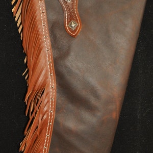May include: Brown leather chaps with fringe detail and silver accents. The chaps have a decorative stitching pattern and a belt loop.