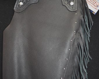 May include: Black leather chaps featuring a textured surface, long fringe along the side, and a yoke with intricate floral tooling. The garment includes silver-toned metal studs, a decorative silver buckle, and a small attached product tag.