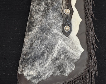 May include: Brown leather chaps with a white and black cowhide pattern. The chaps have a fringe trim and decorative silver conchos.