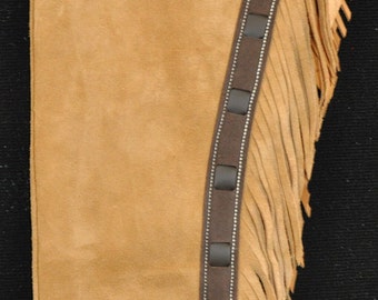 Può includere: Un paio di chaps in pelle marrone con dettagli a frangia e un cinturino in pelle marrone con accenti argentati.