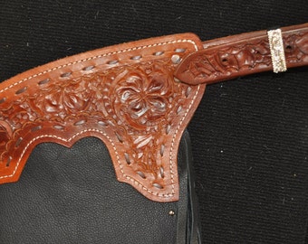 May include: Brown leather chaps with a floral tooled design and a silver buckle. The chaps have a white stitching detail.