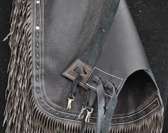 May include: Black leather chaps with fringe and a decorative design. The chaps have a belt loop and a buckle closure.