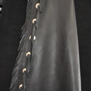 May include: Black leather chaps with decorative silver-tone snaps and fringe along the side. The top portion features embossed floral designs. A tag with an American flag is visible.