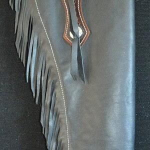 May include: Black leather chaps with brown tooled leather accents and fringe detailing. Features include a decorative brown leather yoke with floral tooling, white stitching, and silver-toned hardware. A classic Western wear item.
