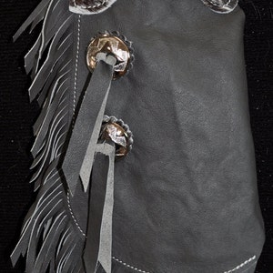 May include: Gray leather chaps with fringe and a cowhide patch. The chaps have two silver concho buttons with leather straps.
