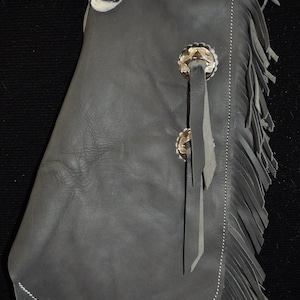 May include: Gray leather chaps with fringe and a white and black cowhide patch. The chaps have two decorative silver conchos with leather tassels.
