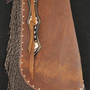 May include: Brown leather chaps with fringe and white stitching. The chaps have a black leather panel with silver accents and a brown leather strap.