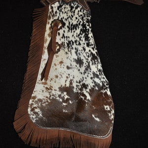 May include: Brown leather chaps with a white and brown spotted pattern and fringe trim. The chaps have a belt loop and two decorative metal buckles.