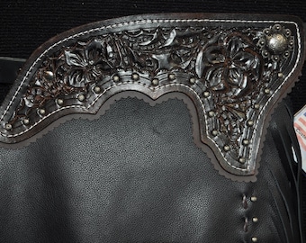 May include: Close-up of a black leather chaps with intricate floral tooling on the upper portion. The tooling is dark brown with silver-colored studs and white stitching. A silver concho is visible.