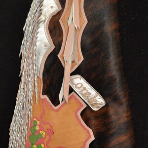 May include: Hand-tooled leather chaps with a brown and black cowhide design. The chaps feature silver fringe, cactus and floral accents, and the name "Corelei" on a silver tag. The chaps have a leather belt.