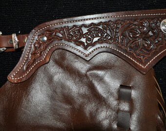 Può includere: Chaps in pelle marrone con elaborate lavorazioni floreali e una concho argentata. I chaps hanno un passante per la cintura e un dettaglio di cucitura bianca.
