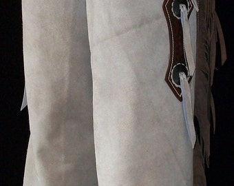 May include: A pair of white leather chaps with brown leather trim and fringe. The chaps have a decorative design on the side with white leather straps and metal accents.