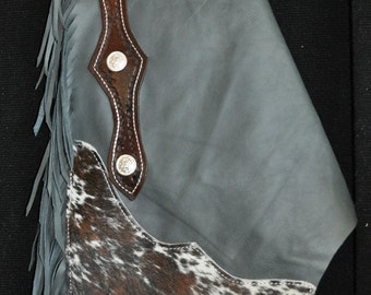 May include: Gray leather chaps with brown leather accents and fringe. The chaps have a cowhide patch on the lower leg and silver conchos.