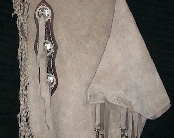 Può includere: Un paio di chaps in pelle marrone con frange e un dettaglio di cucitura decorativo. I chaps hanno una cinghia in pelle marrone con una fibbia.