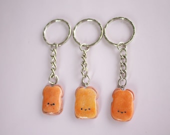 Puede incluir: Tres llaveros con un diseño de tostadas de dibujos animados con caras sonrientes. Cada llavero está hecho de arcilla polimérica de color marrón claro y naranja y tiene una cadena y un anillo de metal plateado.