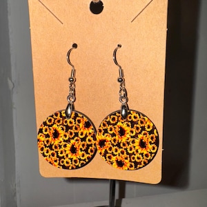 May include: A pair of round wooden earrings with a sunflower pattern. The earrings are yellow, black, and brown. The earrings are hanging from silver hooks.