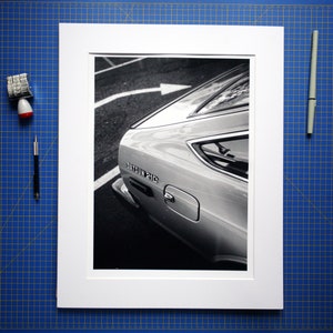 May include: Black and white photograph of a Datsun 210 car's rear. The car is in a white mat, with a blue grid background. A date stamp and pen are on the left and right sides of the mat.