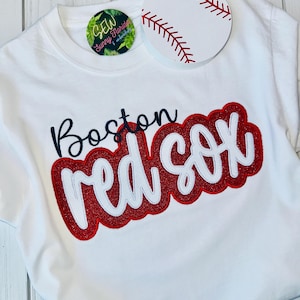 May include: White t-shirt with a red and white "Boston Red Sox" graphic. The graphic is embroidered with glitter. A white baseball with red stitching is on the shirt.