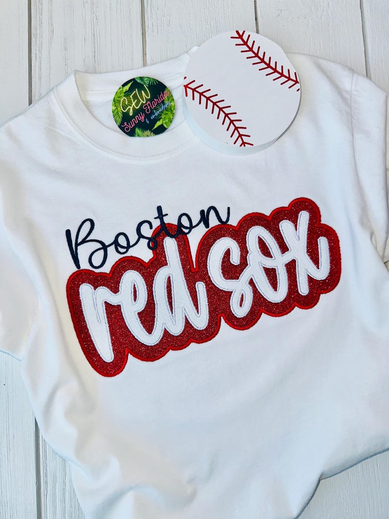 May include: White t-shirt with a red and white "Boston Red Sox" graphic. The graphic is embroidered with glitter. A white baseball with red stitching is on the shirt.