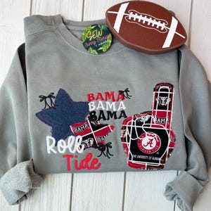 May include: A gray sweatshirt with a star and foam finger design. The star is blue denim, and the foam finger features the University of Alabama logo. The words "Roll Tide" and "Bama" are also displayed. A brown football-shaped item is on top.