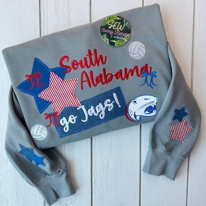 May include: Gray sweatshirt with embroidered patches. Features the words "South Alabama" in red, a blue and red star, a volleyball, and the text "go Jags!" in white on a blue patch. A panther logo is also visible.