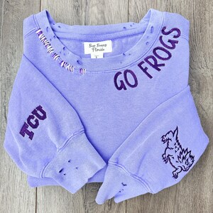 May include: Purple sweatshirt with distressed detailing and embroidered text "GO FROGS" on the neckline and "TCU" on the sleeve. The sweatshirt also features an embroidered frog on the right sleeve.