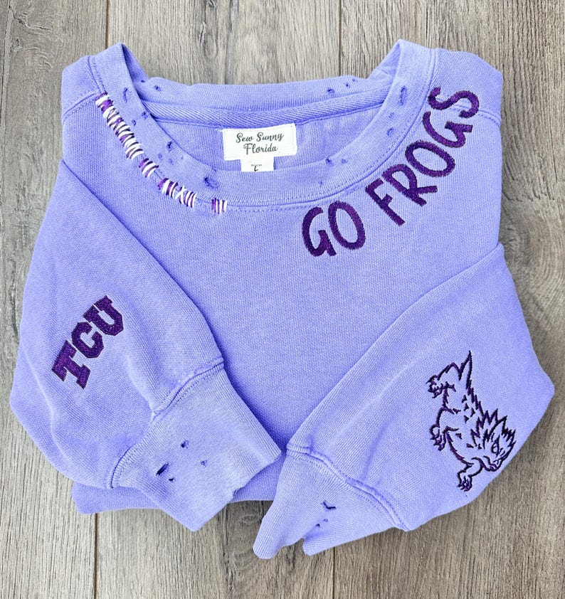 May include: Purple sweatshirt with distressed detailing and embroidered text "GO FROGS" on the neckline and "TCU" on the sleeve. The sweatshirt also features an embroidered frog on the right sleeve.
