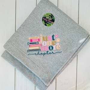 May include: A folded gray blanket with the embroidered text "just one more chapter" and a stack of books in pink, gold, and teal. A circular logo with text and foliage is in the upper corner.