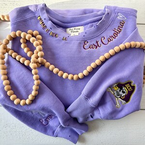 May include: Purple sweatshirt with distressed details, featuring embroidered text "East Carolina" and a skull and crossbones logo with "ECU" on the sleeve.