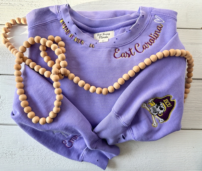 May include: Purple sweatshirt with distressed details, featuring embroidered text "East Carolina" and a skull and crossbones logo with "ECU" on the sleeve.