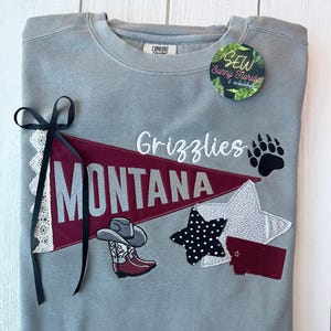 May include: Gray sweatshirt with a Montana Grizzlies design. Features a maroon pennant with "MONTANA" in silver, a cowboy boot and hat, and a paw print. Includes a black bow with lace trim and a "SEW Sunny Florida" tag.