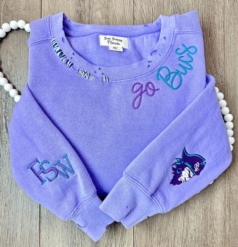 May include: A lavender sweatshirt with the text "go Bucs" embroidered on the front in purple and teal thread. The left sleeve has "FSW" embroidered in purple and teal thread. The right sleeve has a teal and purple embroidered graphic of a fish with a shark fin.