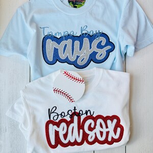 May include: Two t-shirts with embroidered baseball team logos. The blue shirt has "Tampa Bay Rays" in blue and silver glitter. The white shirt has "Boston Red Sox" in red and white glitter.