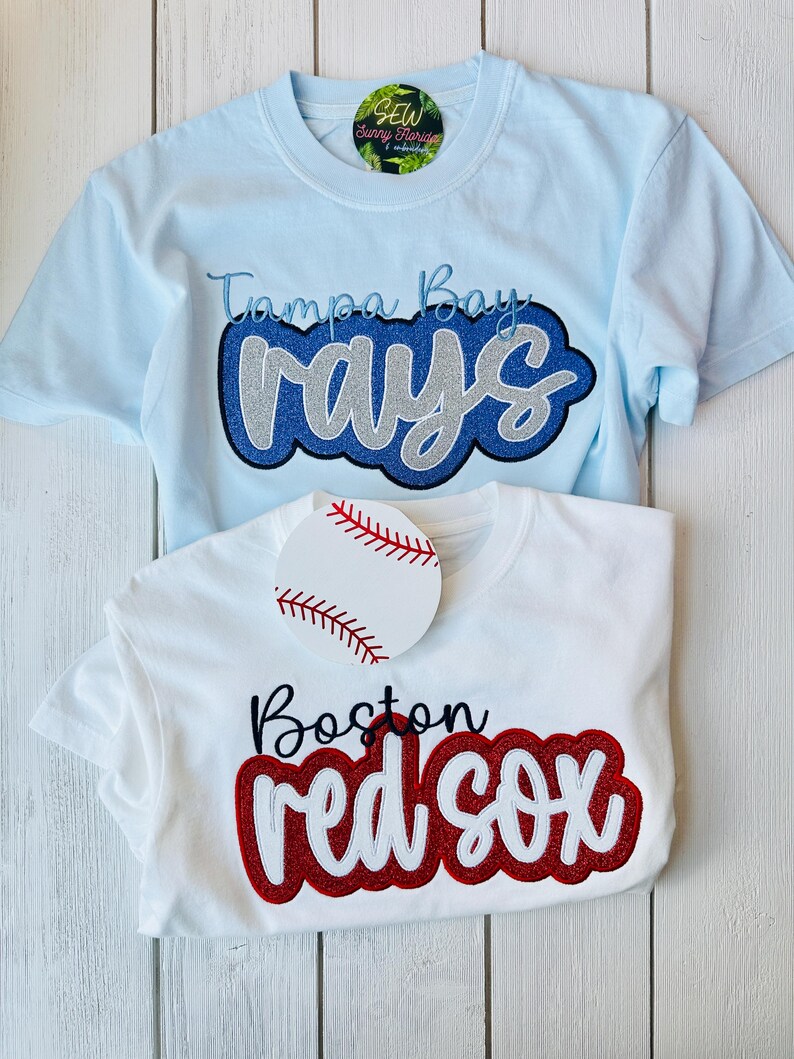 May include: Two t-shirts with embroidered baseball team logos. The blue shirt has "Tampa Bay Rays" in blue and silver glitter. The white shirt has "Boston Red Sox" in red and white glitter.