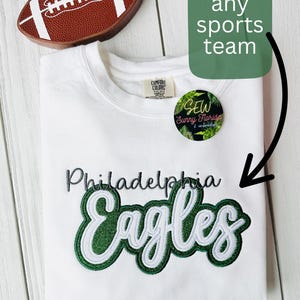 May include: White sweatshirt with the Philadelphia Eagles logo in green and white. A brown football-shaped item is at the top left. The text "Choose any sports team" is in a green rectangle.
