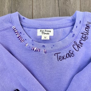 May include: Purple sweatshirt with a distressed neckline and "Texas Christian" embroidered in black on the right shoulder. The sweatshirt has a white tag that reads "Sew Sunny Florida" and a size "L" tag.