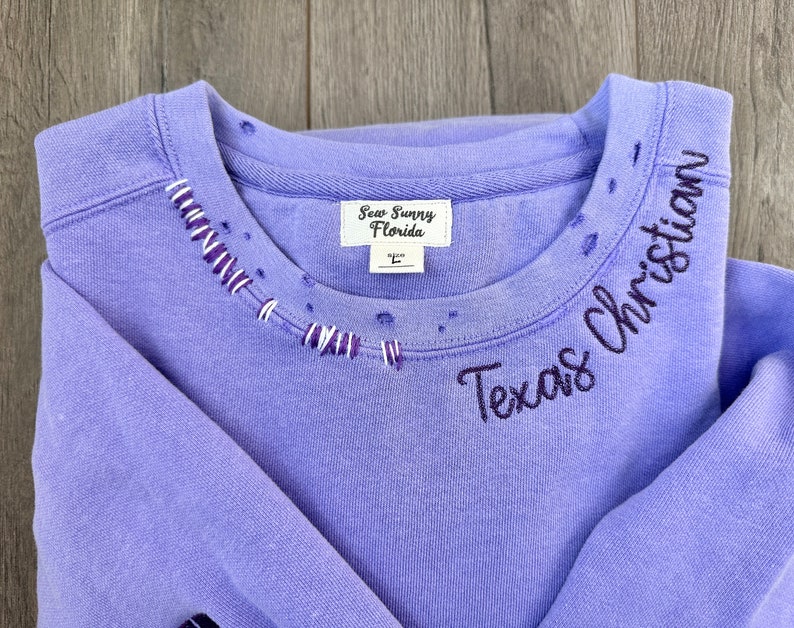 May include: Purple sweatshirt with a distressed neckline and "Texas Christian" embroidered in black on the right shoulder. The sweatshirt has a white tag that reads "Sew Sunny Florida" and a size "L" tag.
