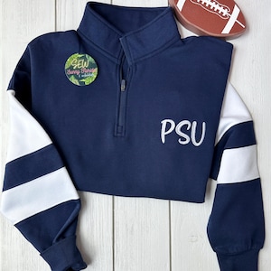 May include: Navy blue quarter-zip sweatshirt with white striped sleeves and the embroidered letters "PSU". A small round pin with the text "SEW Sunny Florida" is attached. A decorative football is placed on top.