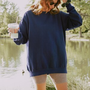May include: A woman wearing a navy blue oversized sweatshirt with a light brown pair of shorts. She is holding a clear plastic cup with a straw.