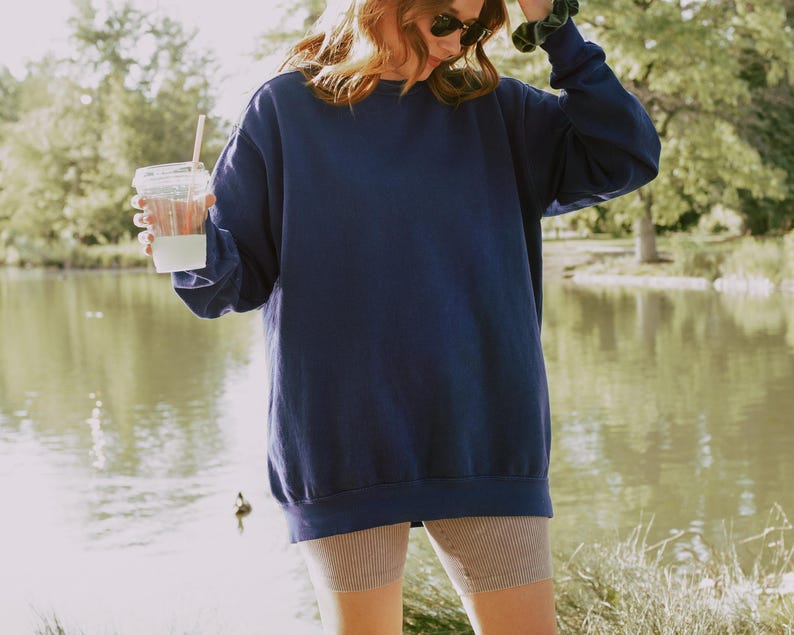 May include: A woman wearing a navy blue oversized sweatshirt with a light brown pair of shorts. She is holding a clear plastic cup with a straw.