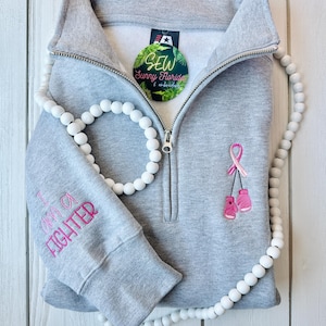 May include: Gray sweatshirt with a pink ribbon and boxing gloves embroidered on the front. The left sleeve has the text "I am a fighter" embroidered in pink. A white bead necklace and a round tag with the text "SEW Sunny Florida & embroidery" are also visible.