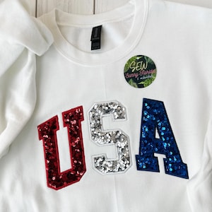 May include: White sweatshirt with the letters "USA" appliqued on the front. The "U" is red, the "S" is silver, and the "A" is blue, all with sequin detailing. A circular sticker with text is on the upper right.