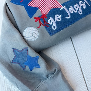 May include: A gray sweatshirt with embroidered stars, a volleyball, and the text "So Alabama go Jags!". The design includes blue, red, and white accents. The sleeve features two blue stars.