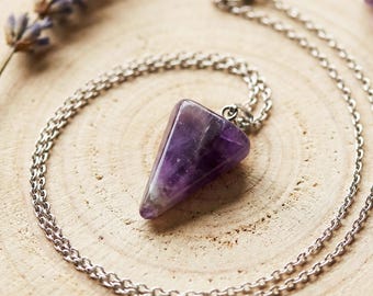 May include: A silver chain necklace with a triangular, purple amethyst pendant. The necklace is displayed on a wooden surface, accompanied by sprigs of lavender and several raw amethyst crystals. The pendant is approximately 1 inch in length.